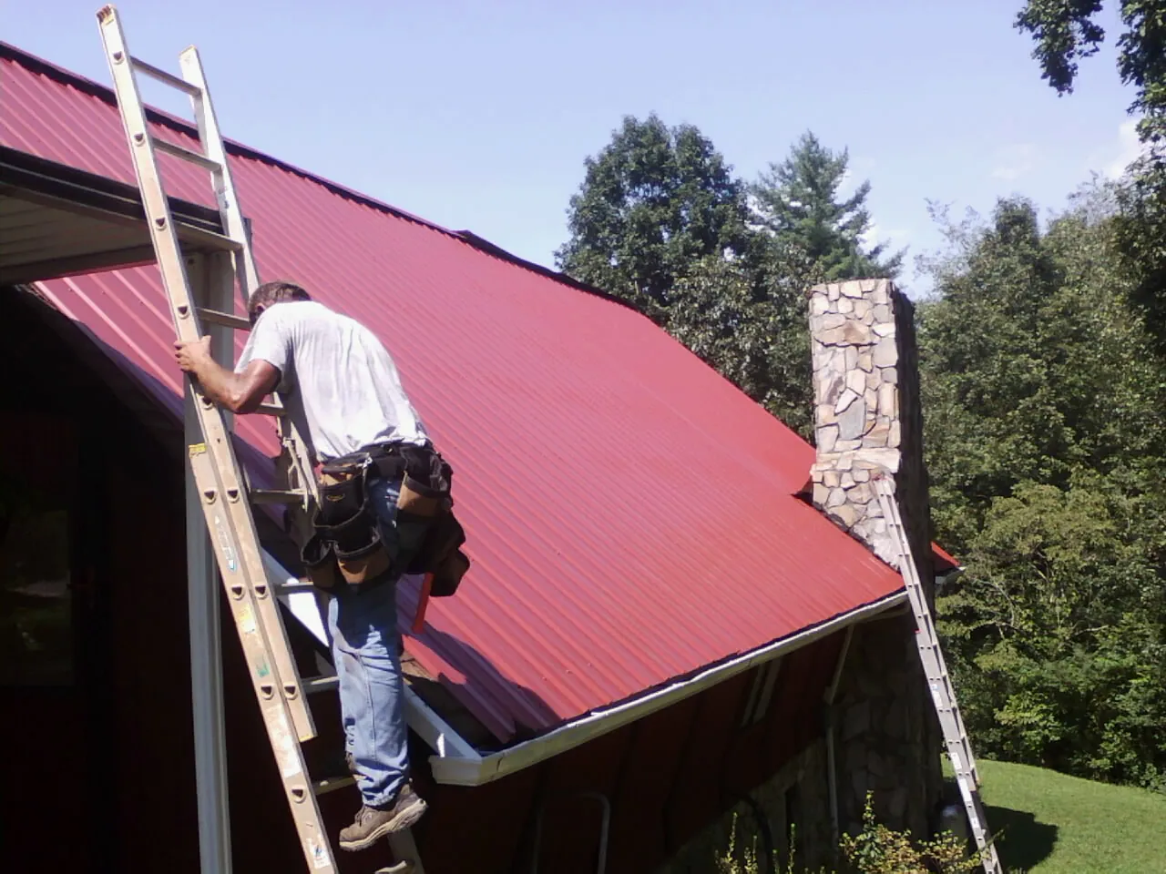 Roofer on a ladder beside a red metal roof and stone chimney — Soffit & Fascia Repair by Roofing Brothers in Mehoopany